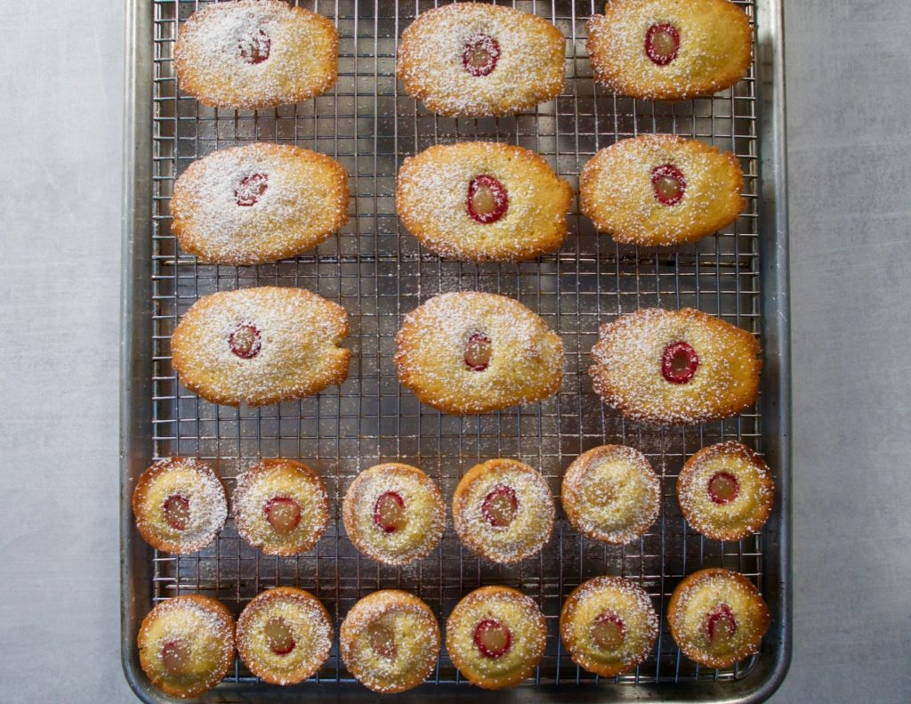 Lemon curd and raspberry filled madeleines resting on a cooling rack.