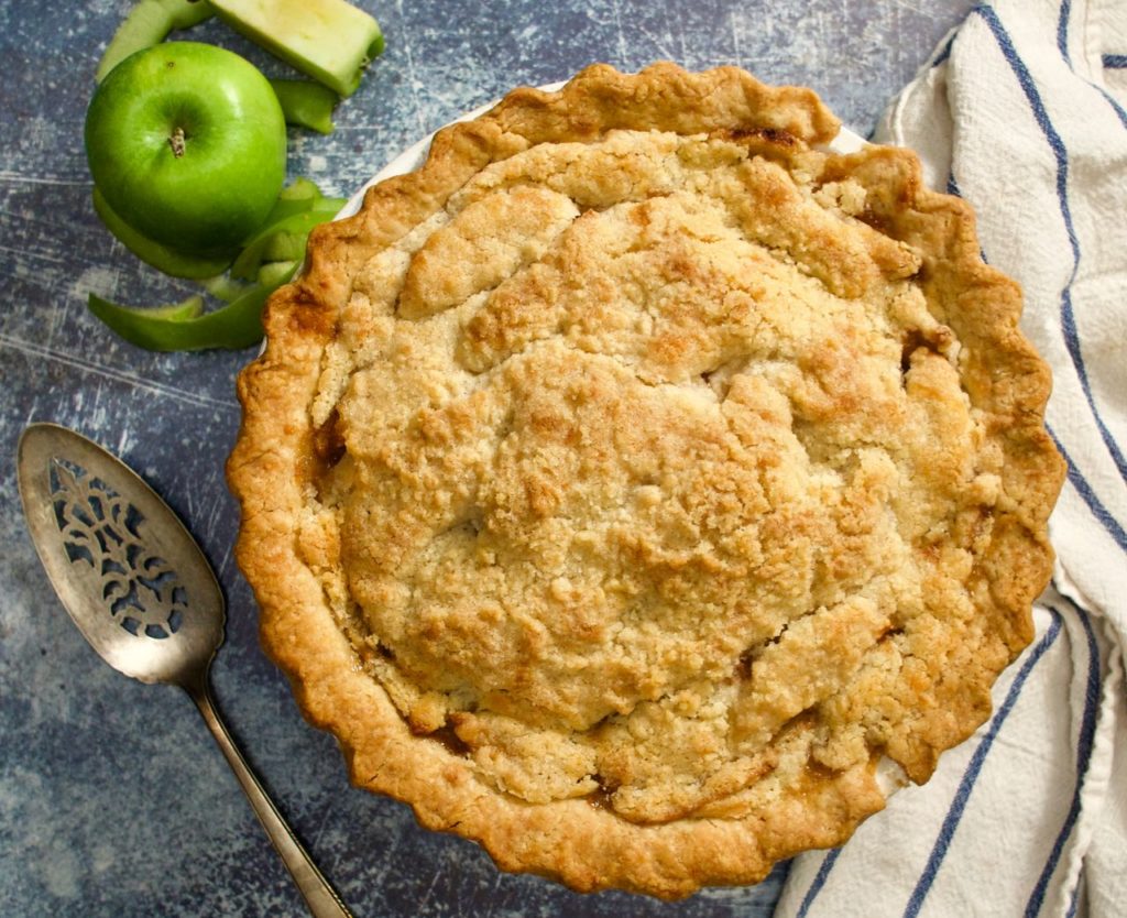 Whole apple pie with pie server, apples and striped towel.