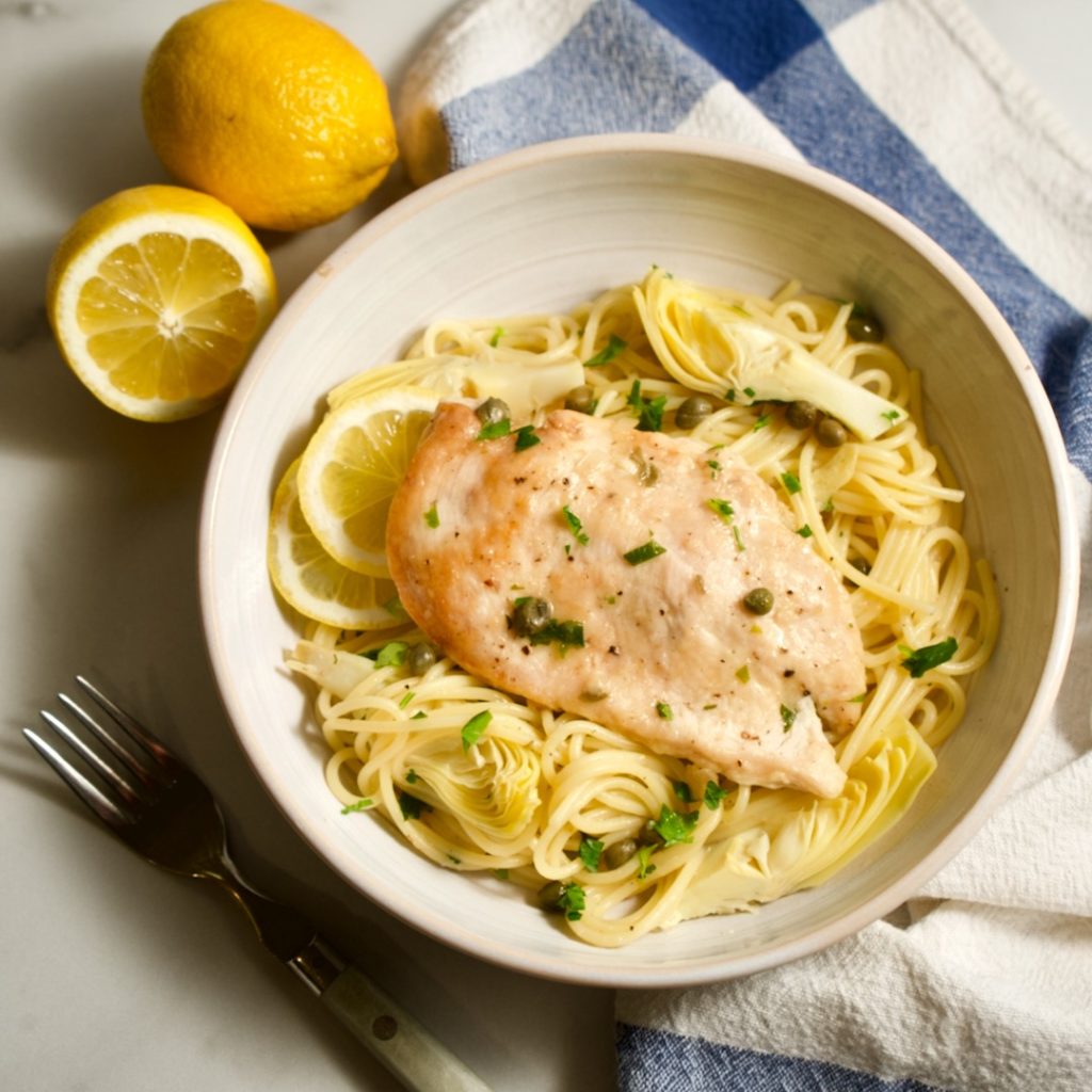 Chicken piccata pasta in bowl with fork, lemons and napkin.