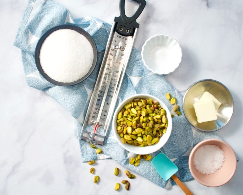 Bowl of sugar, candy thermometer, pistachios, butter and salt with spatula.