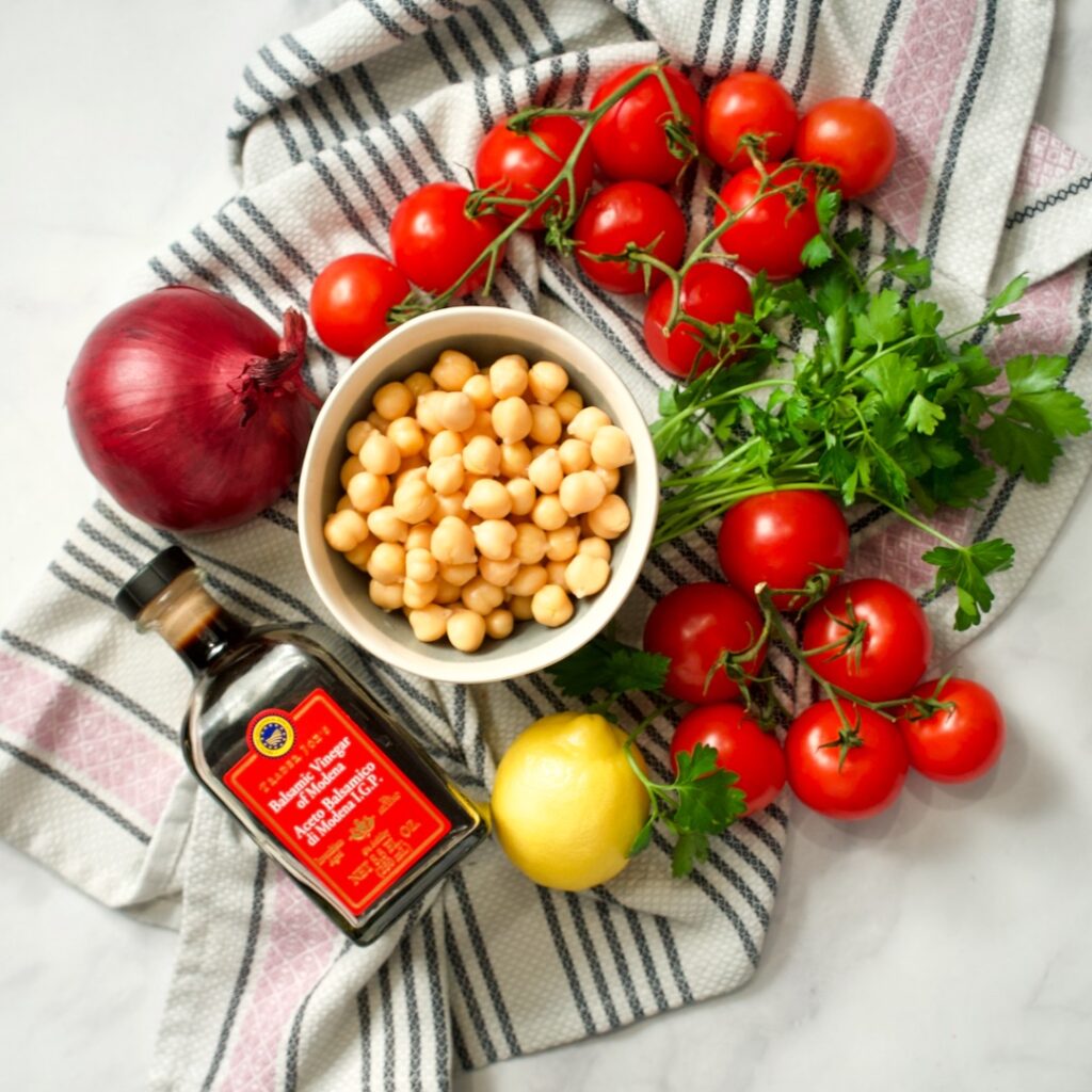 Vine tomatoes, canned chickpeas, parsley, lemon, red onion and a bottle of balsamic on kitchen towel.