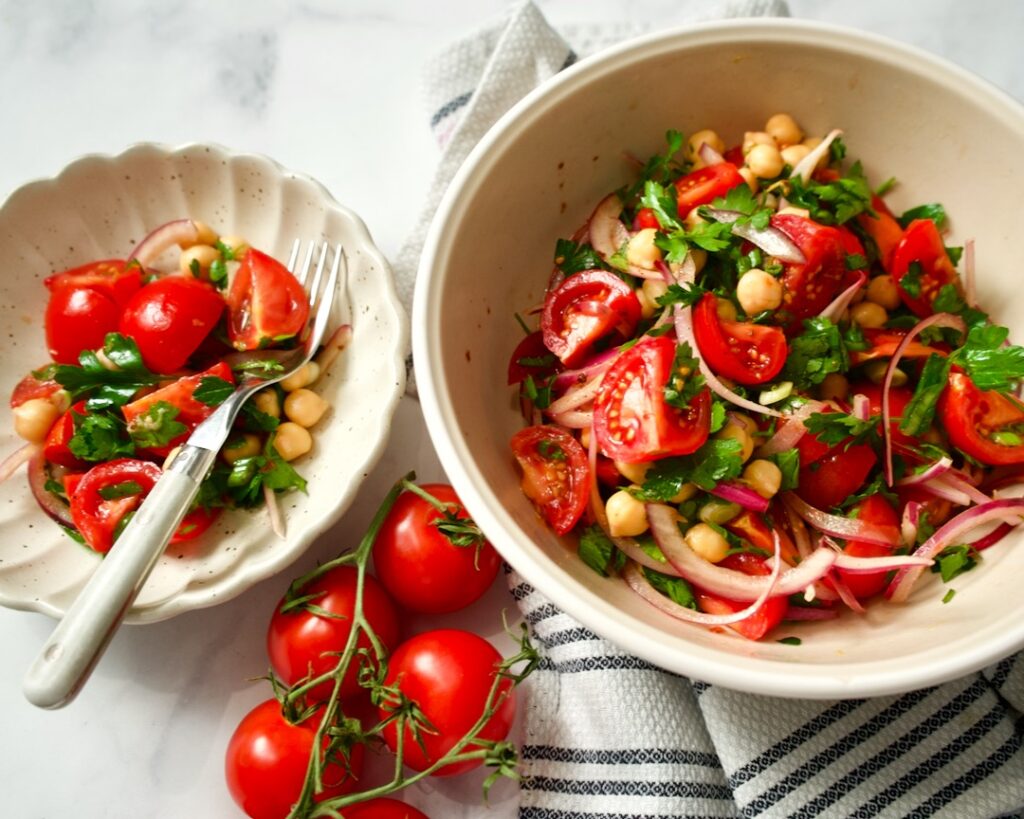 Large salad bowl on towel next to vine tomatoes. Small serving bowl filled with salad.