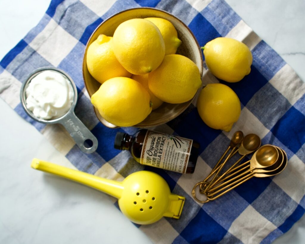 Bowl of lemons, greek yogurt in measuring cup, vanilla bottle, measuring spoons and a lemon squeezer in blue towel.