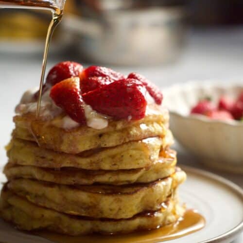Johnny cakes on a plate with strawberries in a bowl behind. Maple syrup poured on top.